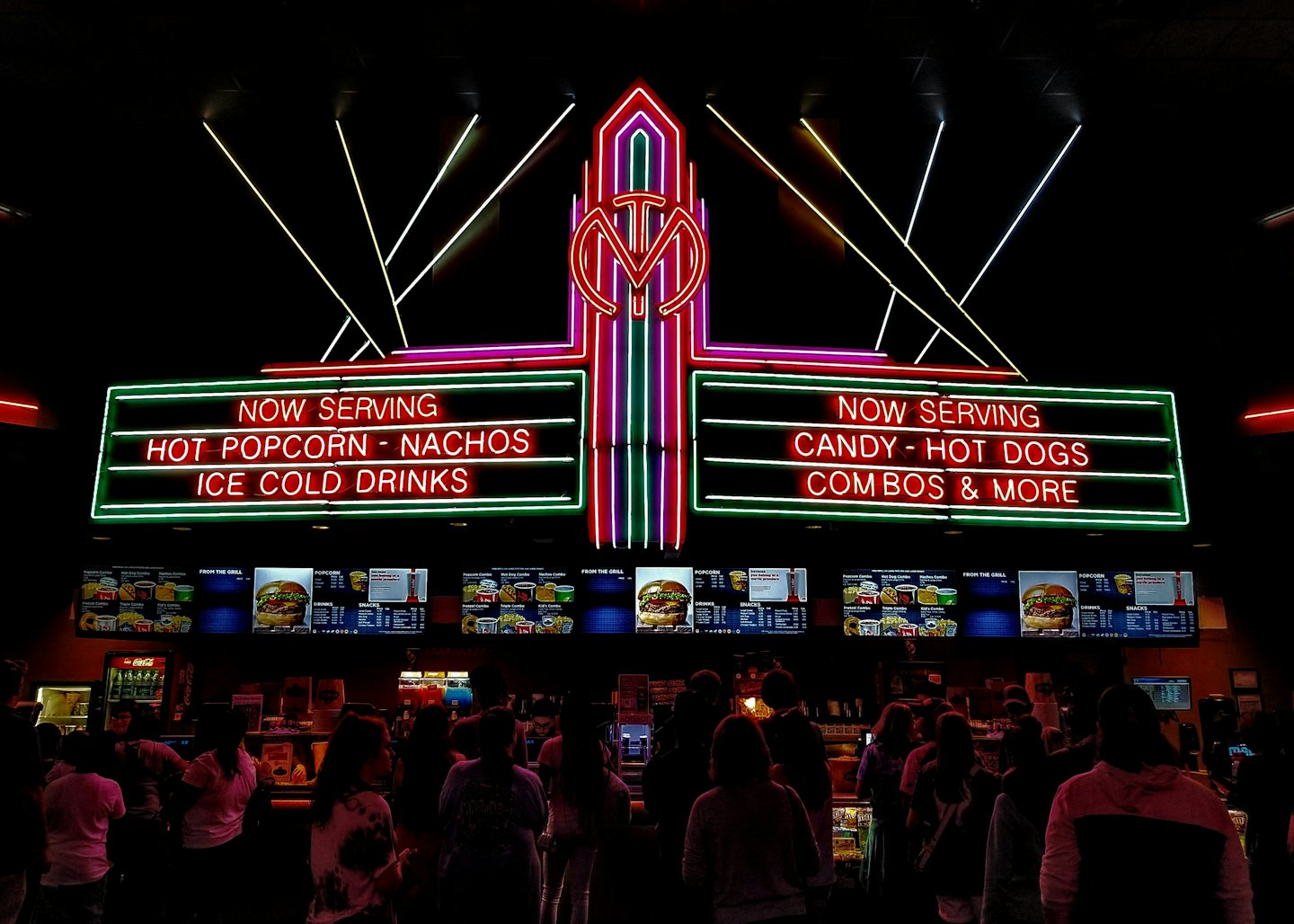Persone in fila al banco del cibo di un cinema con insegne al neon.